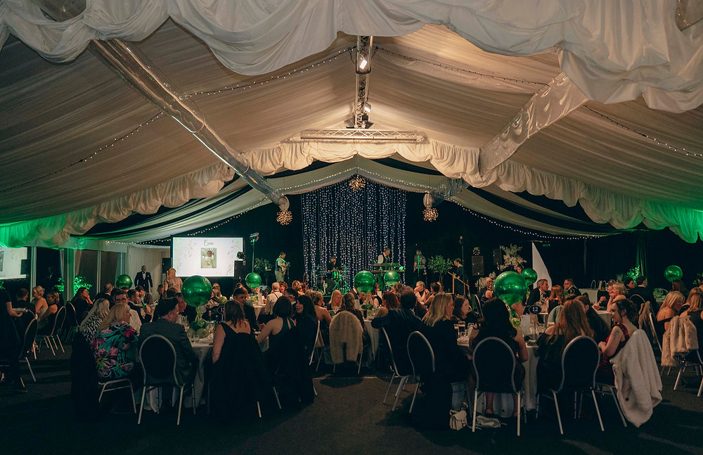 Guests seated at round tables in a marquee at the Team Evie black tie ball, decorated with green balloons and fairy lights